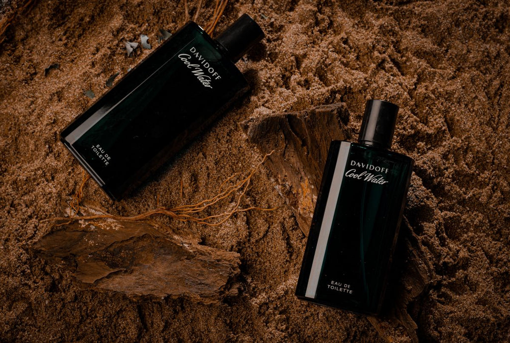 Two dark rectangular fragrance bottles resting on sand with pieces of driftwood